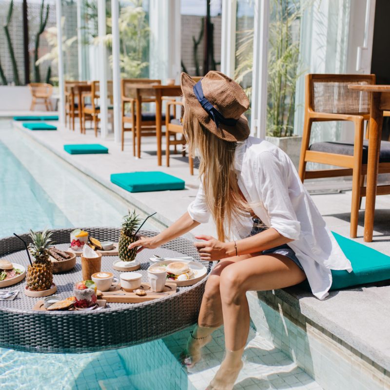 Slim girl in elegant brown hat eating juicy fruits at resort cafe. Graceful european woman in white shirt relaxing with cocktail and food in pool.